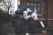 A trash can full of garbage in front of a brick building. A trash can full of garbage in front of a brick building.